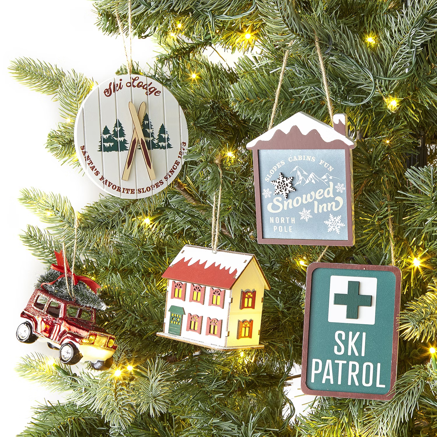 A collection of Christmas tree ornaments including a ski lodge sign, a Snowed Inn cabin plaque, a small house, a red car with a tree on top, and a Ski Patrol badge.