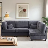 A gray corduroy sectional sofa with two cushions is set in a modern living room. The decor includes a framed abstract art piece, a floor lamp, a wood coffee table with books and a throw, a light area rug, and a potted plant near the curtains.