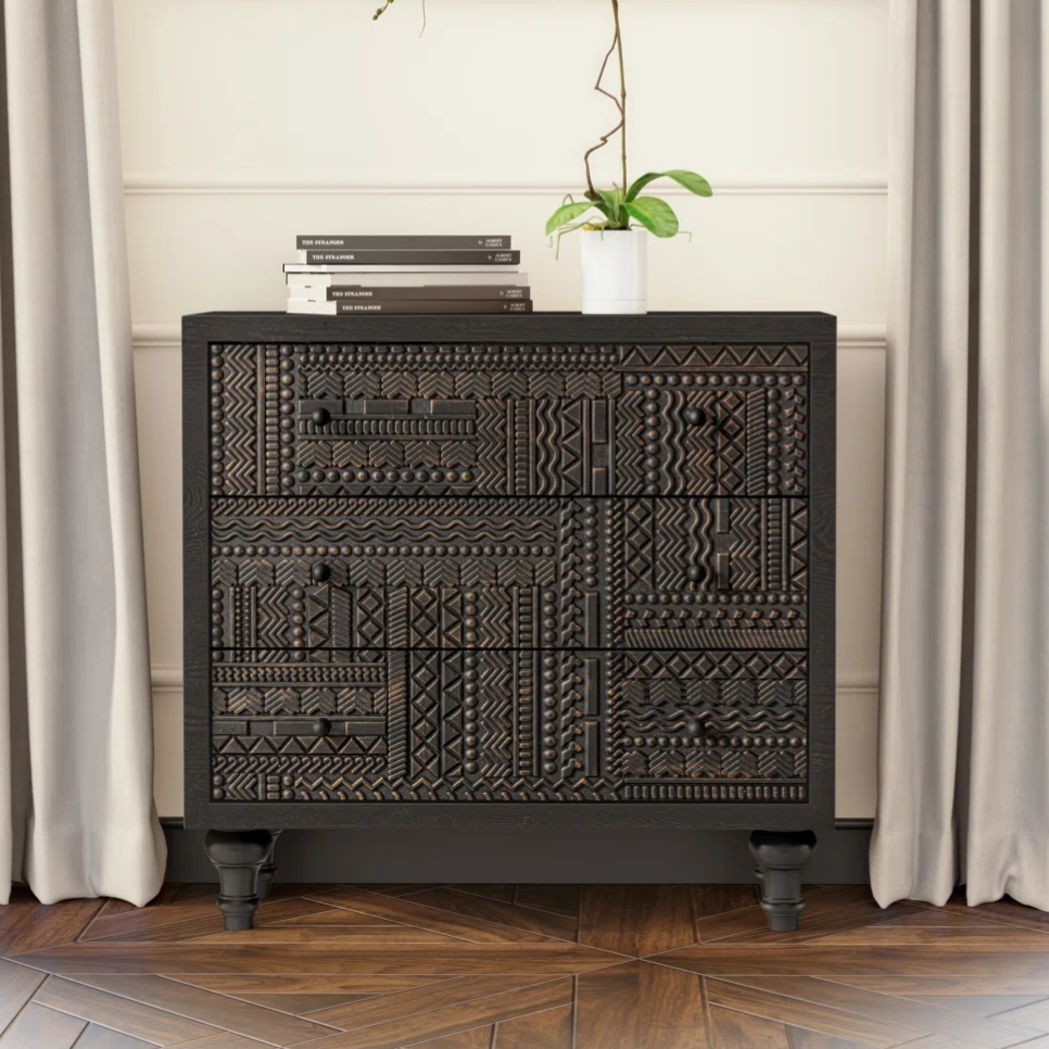 A black, intricately carved dresser stands against a wall with a modern painting above it, flanked by taupe curtains. A white orchid in a small pot and stacked books adorn the dresser.