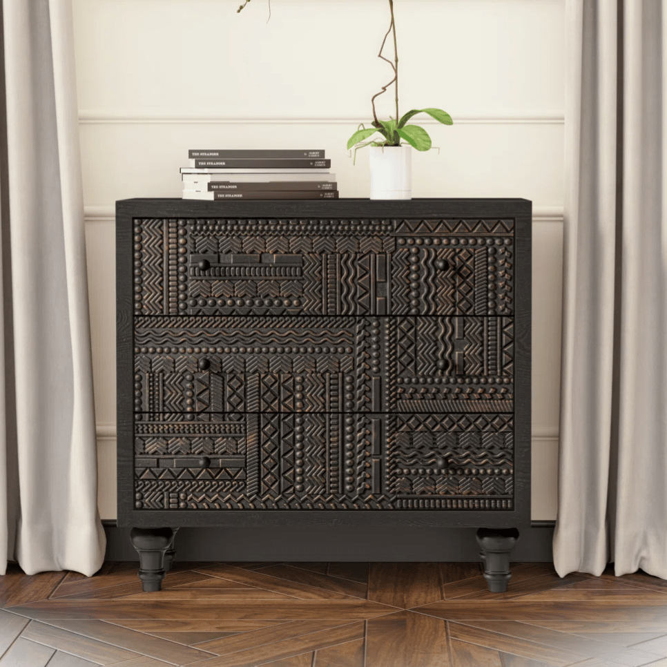 A black, intricately carved dresser stands against a wall with a modern painting above it, flanked by taupe curtains. A white orchid in a small pot and stacked books adorn the dresser.