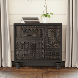 A black, intricately carved dresser stands against a wall with a modern painting above it, flanked by taupe curtains. A white orchid in a small pot and stacked books adorn the dresser.