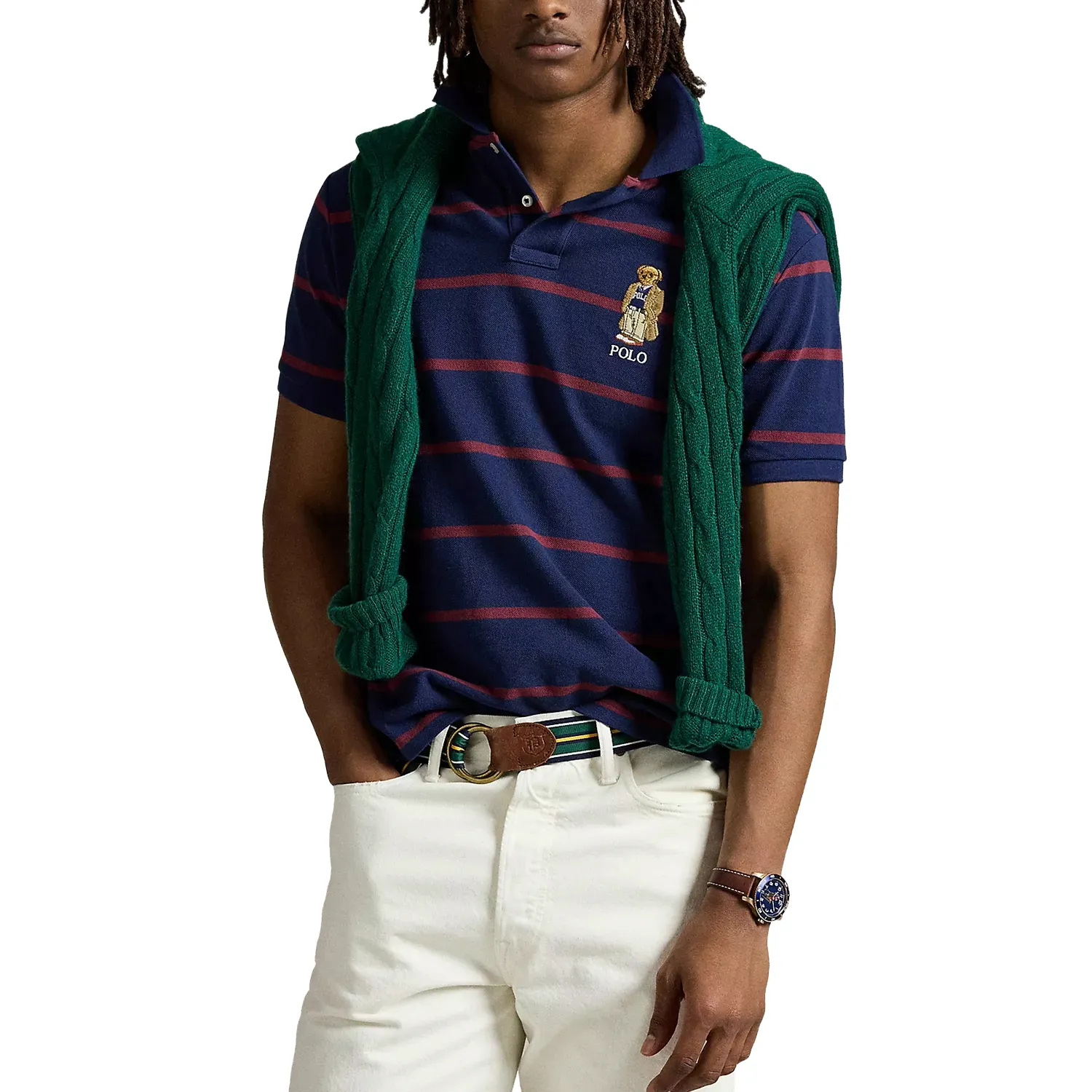 Man wearing a navy and red striped Polo shirt with a bear logo, cream pants, a green sweater over shoulders, brown belt, and a watch.