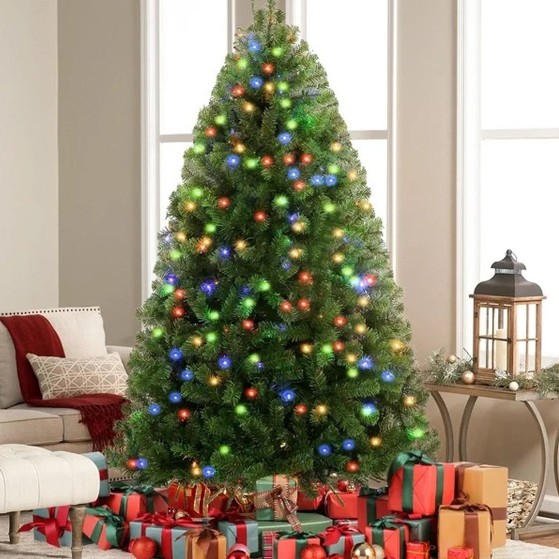 A decorated Christmas tree with multicolored lights is surrounded by wrapped gifts. There's a cozy seating area with a sofa, ottoman, and lantern on a small table.