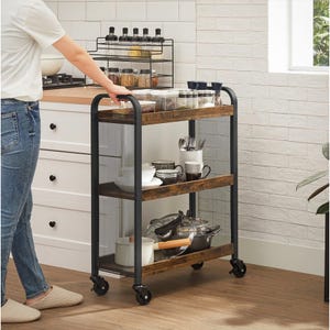 A kitchen cart with three shelves holds dishes, utensils, and pots, with spices and seasonings on the countertop. The cart has a metal frame with wooden shelves and wheels for easy mobility.