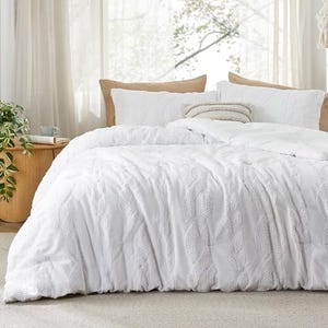 A queen-size tufted comforter set with a textured white design, accompanied by two brown and two white matching pillows, displayed on a bed in a bright room.
