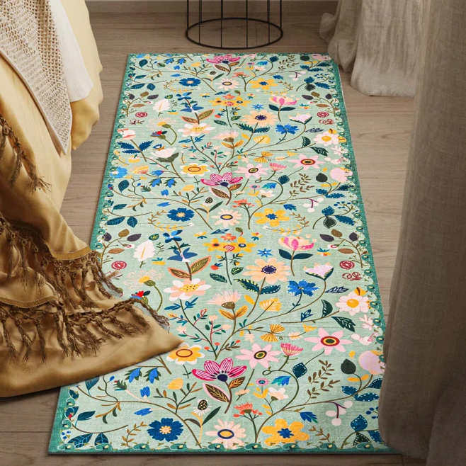A rectangular floral rug with a colorful pattern of various flowers and leaves on a light background lies on a wooden floor beside a bed draped with a fringed blanket.