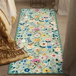 A rectangular floral rug with a colorful pattern of various flowers and leaves on a light background lies on a wooden floor beside a bed draped with a fringed blanket.