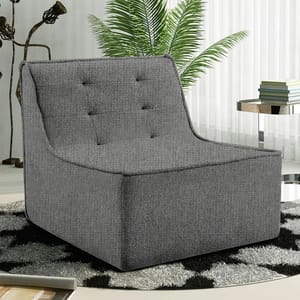 A modern, gray fabric armchair with tufted backrest placed on a black and white patterned rug, alongside a small stack of books on a round side table, with a potted plant and sheer curtains in the background.