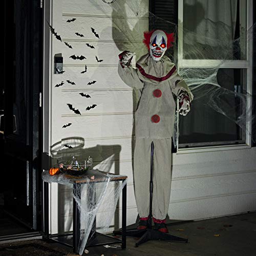A standing clown animatronic with a menacing expression, red hair, and a costume with red accents is positioned next to a small table with Halloween decorations under a porch with bat silhouettes.