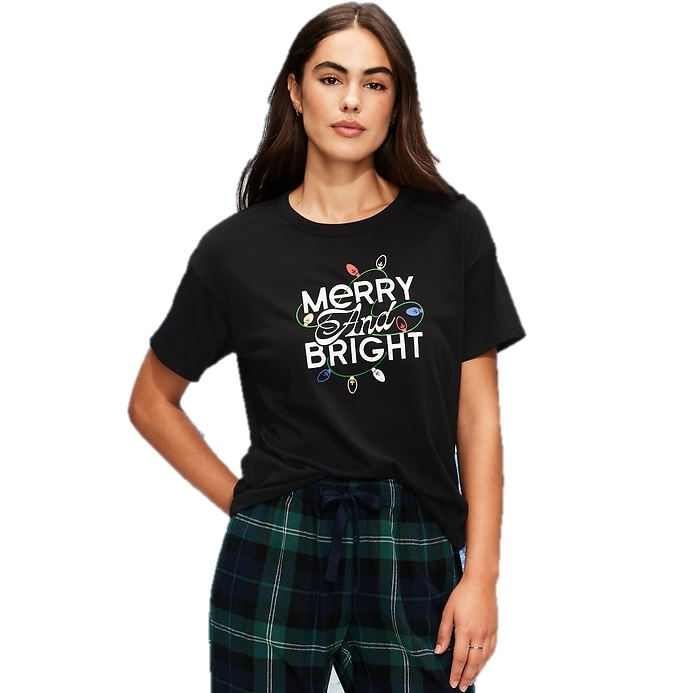 A woman wears a black T-shirt with the text \“Merry And Bright\“ accompanied by colorful Christmas lights, paired with dark green plaid pants.