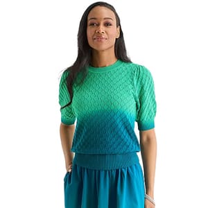 A woman is wearing a short-sleeve knit top with a braided pattern in a gradient from green to blue, paired with a matching blue skirt.