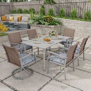 Outdoor dining set: six wicker chairs with cushions around a rectangular table, placed on a stone patio. Decor includes plates, glasses, and a floral centerpiece. Background features a seating area with colorful cushions and surrounding greenery.