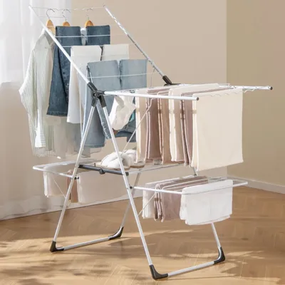 A white metal drying rack holds various clothes and towels in a well-lit room. Nearby, a grey laundry basket with handles contains a folded cloth.