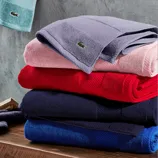 Stack of colorful towels with a small crocodile logo, displayed on a wooden surface against a textured gray wall. The towels vary in colors: light blue, purple, pink, red, navy, and blue.