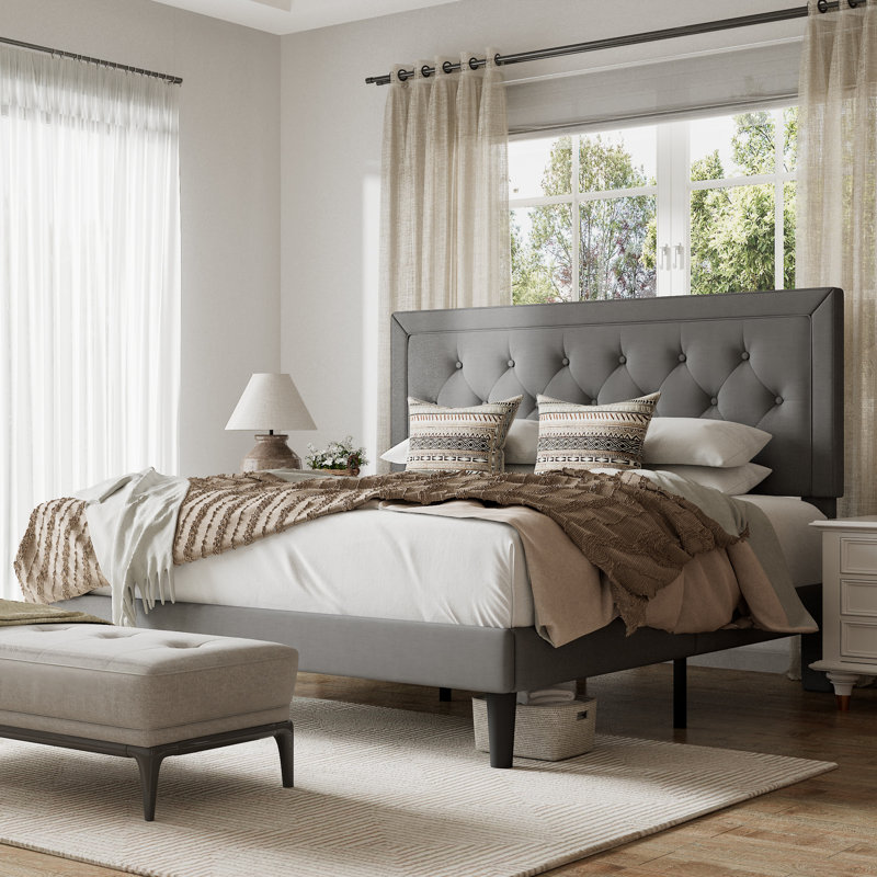 A bedroom features a gray upholstered bed with a tufted headboard, layered bedding in neutral tones, decorative pillows, and a matching bench. A white nightstand with a lamp and sheer curtains accompany the setup for a cozy, elegant look.