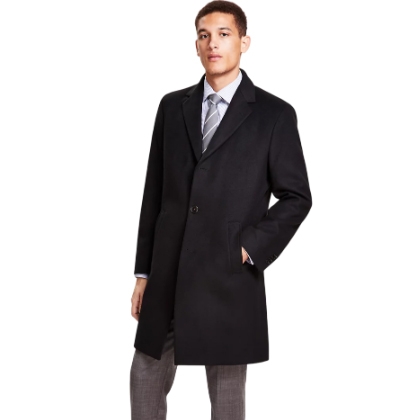 A man is wearing a black, knee-length overcoat paired with a light blue dress shirt, striped tie, and gray trousers.