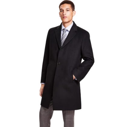 A man is wearing a black, knee-length overcoat paired with a light blue dress shirt, striped tie, and gray trousers.