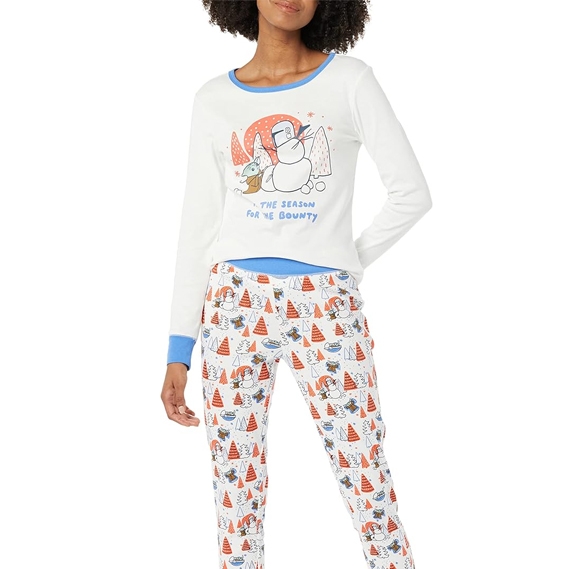 A woman is wearing a white long-sleeve pajama set with blue trim, featuring a snowman and penguin design on the top with the phrase \“The Season for the Bounty,\“ and coordinating pants with a pattern of festive trees and penguins.