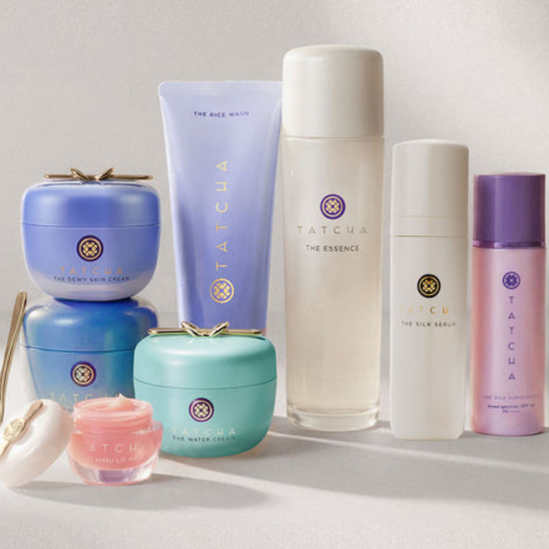 A collection of Tatcha skincare products, including The Rice Wash, The Essence, The Dewy Skin Cream, The Water Cream, The Silk Serum, The Silk Sunscreen, and a lip mask, is displayed.