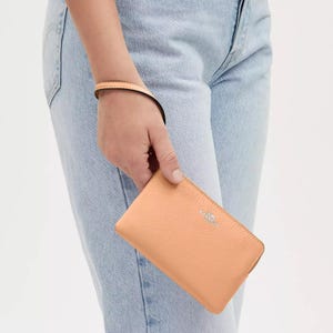 A person is holding a pale pink corner-zip wristlet with a gold-tone logo and matching zipper. The wristlet has a matching strap for easy carrying.