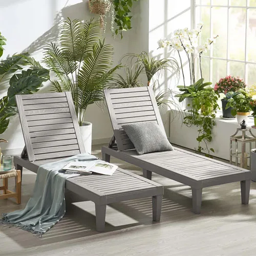 Two gray wooden outdoor lounge chairs with slatted design are placed in a sunlit room with lush green and white plants around, accompanied by a light blue throw blanket and a cushion.