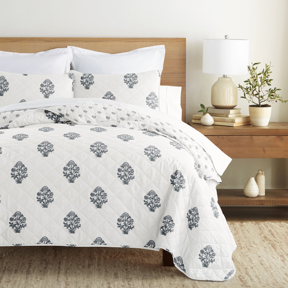 White quilt with a gray floral pattern, accompanied by matching pillows on a wooden bed. A light-colored bedside table holds a lamp, books, and potted plants.