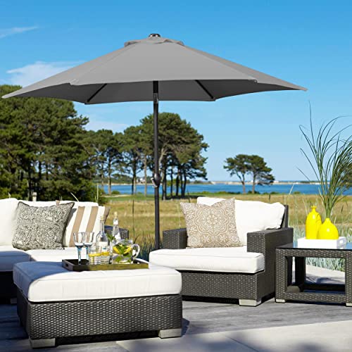 A 7.5-foot patio umbrella in gray provides shade over a seating area with white cushions on wicker furniture, set against a coastal backdrop.