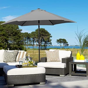 A 7.5-foot patio umbrella in gray provides shade over a seating area with white cushions on wicker furniture, set against a coastal backdrop.
