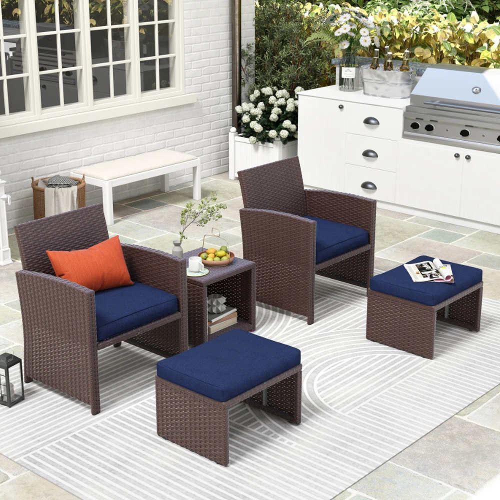 Outdoor patio set with two wicker armchairs, two matching ottomans, and a small side table with blue cushions, set on a striped rug. Nearby is a white outdoor cooking station and potted flowers, creating a modern garden setting.