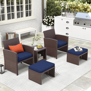 Outdoor patio set with two wicker armchairs, two matching ottomans, and a small side table with blue cushions, set on a striped rug. Nearby is a white outdoor cooking station and potted flowers, creating a modern garden setting.