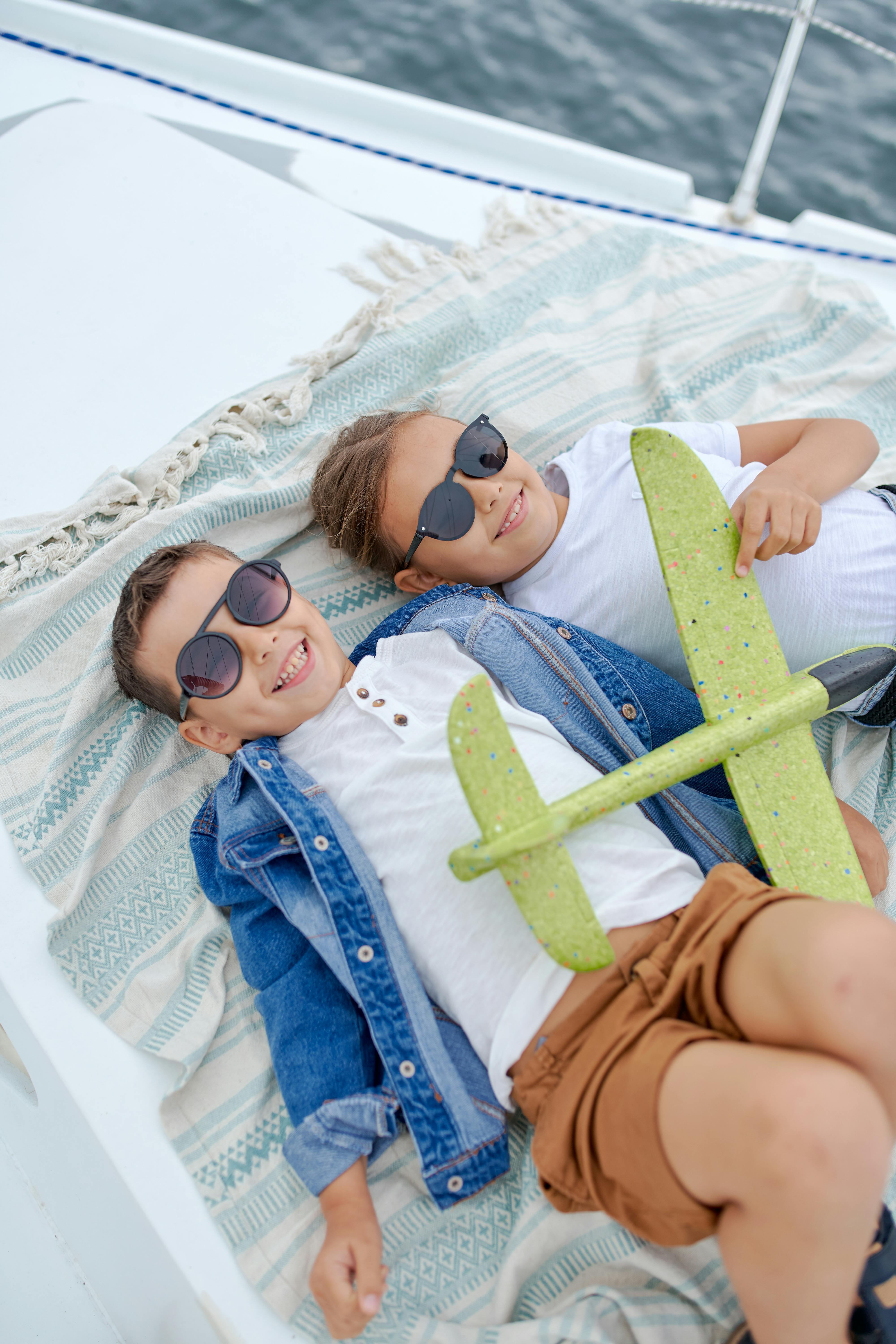 Two smiling children wearing sunglasses and casual clothing, reclining on a boat with a patterned blanket and a toy plane.