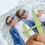 Two smiling children wearing sunglasses and casual clothing, reclining on a boat with a patterned blanket and a toy plane.