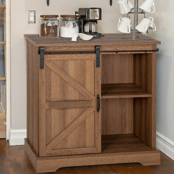A wooden cabinet with a sliding barn door features shelves for storage. On top, there is a coffee maker and jars, and a mug rack holds several white mugs.