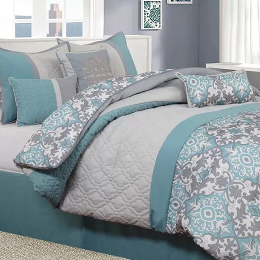 A bedding set featuring a quilted comforter, decorative pillows, and a patterned bedspread in shades of grey and teal with intricate designs.