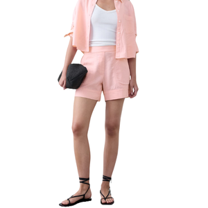 A person wears a light pink button-up shirt, matching shorts, and a white tank top, paired with black strappy sandals and holding a textured black clutch.