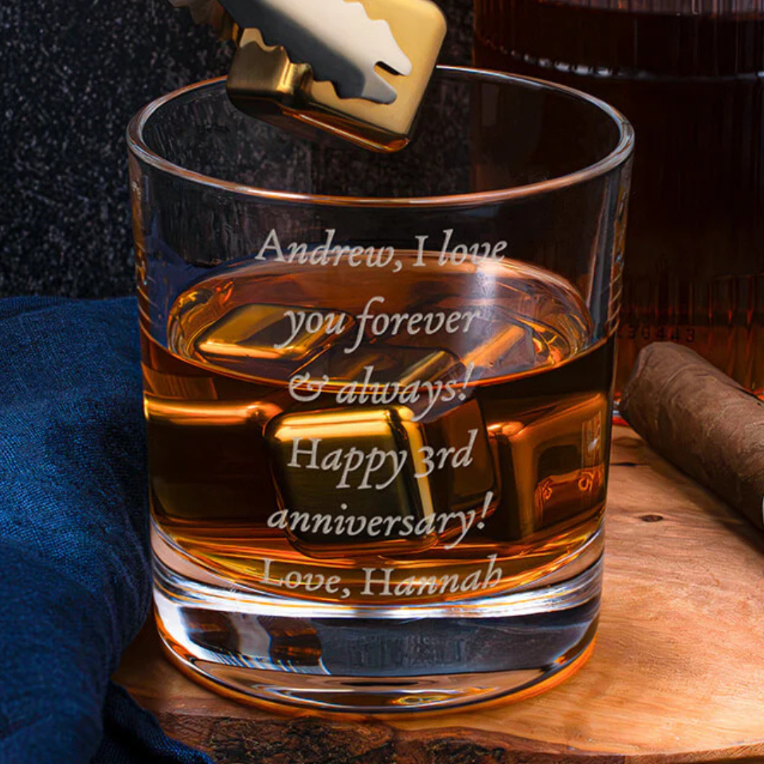A glass with a personalized engraving reads, \“Andrew, I love you forever & always! Happy 3rd anniversary! Love, Hannah,\“ accompanied by whiskey and a cigar on a wooden surface.