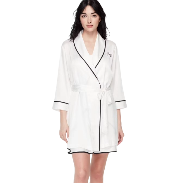 A woman is modeling a white robe with black trim and a waist tie. The robe features an embroidered name on the chest.