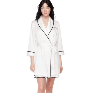 A woman is modeling a white robe with black trim and a waist tie. The robe features an embroidered name on the chest.