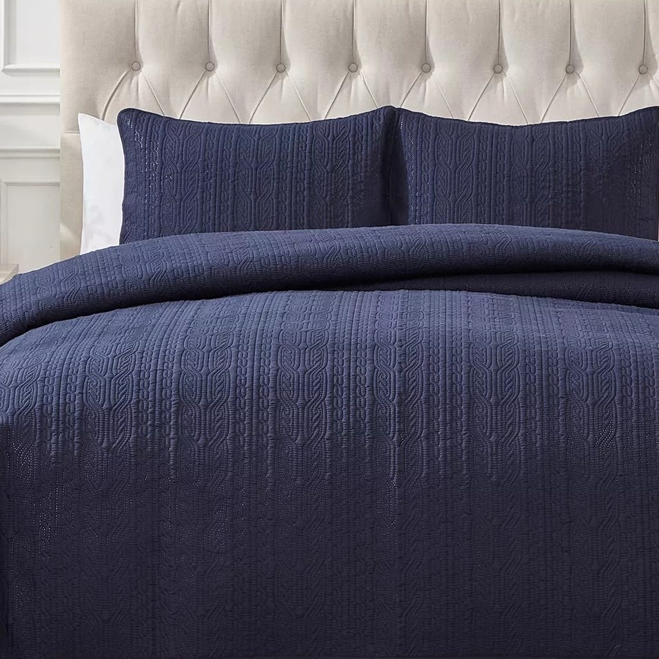 A navy blue quilted bedspread and matching pillow shams with a textured, geometric pattern are displayed on a bed with a tufted headboard.