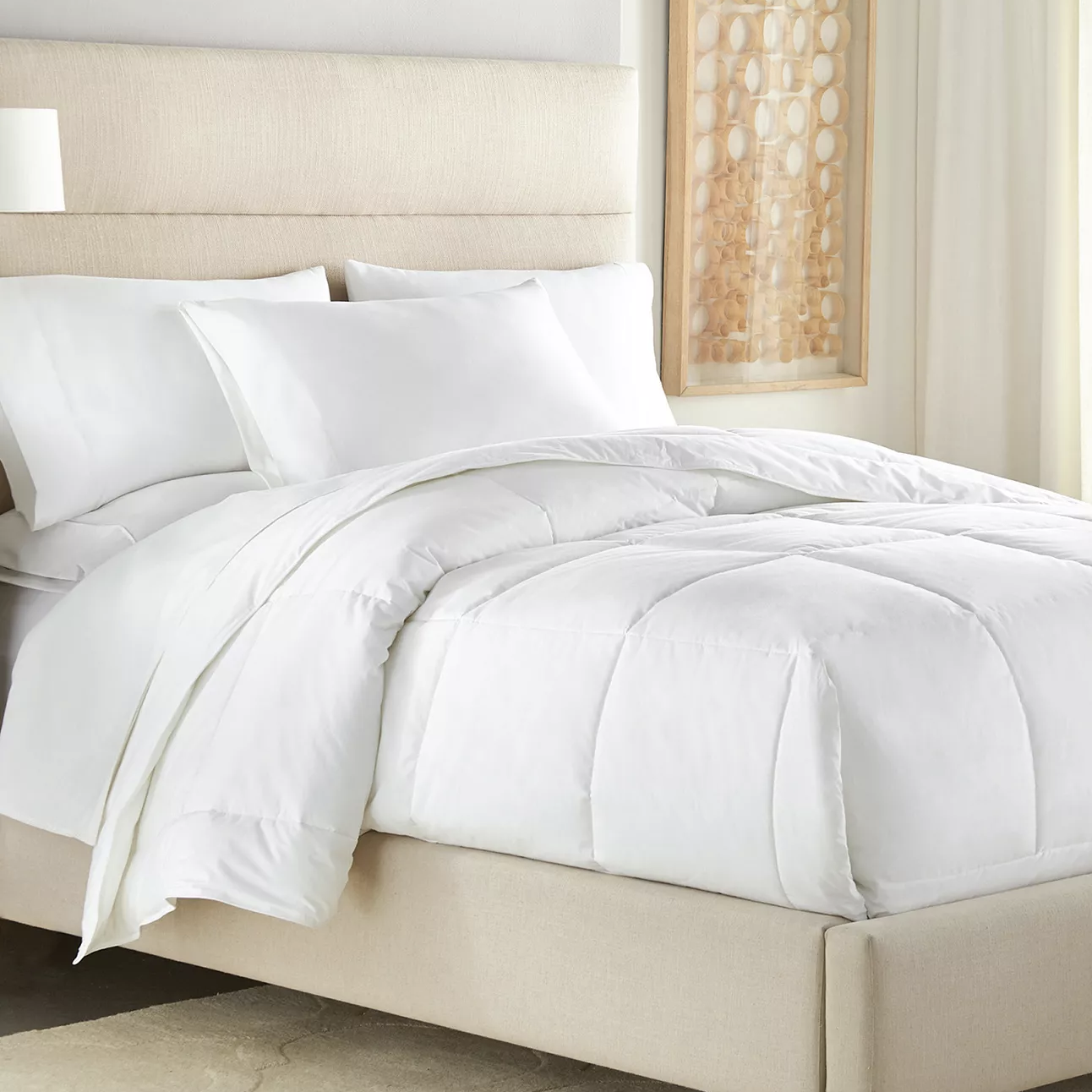 A neatly made bed features a plump white comforter with two matching white pillows, set against a neutral headboard. A framed artwork with circular patterns hangs beside the bed.