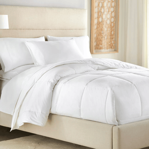A neatly made bed features a plump white comforter with two matching white pillows, set against a neutral headboard. A framed artwork with circular patterns hangs beside the bed.