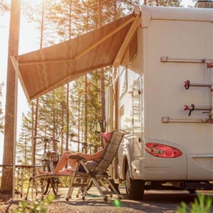 An RV with an awning extended is parked in a forested area. A person relaxes on a reclining chair next to a bicycle.