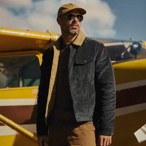A man stands by a yellow airplane wearing a black corduroy and shearling jacket, a brown cap, sunglasses, and brown pants.