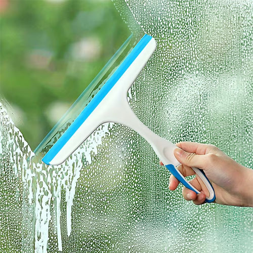 A hand is using a white and blue squeegee to clean a glass window, effectively removing water and leaving a clear path through the droplets.