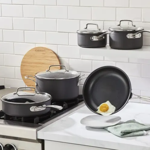 Nonstick cookware set with pots, pans, and lids on a kitchen stove and counter; a fried egg slides off a frying pan, demonstrating nonstick capabilities.