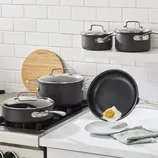 Nonstick cookware set with pots, pans, and lids on a kitchen stove and counter; a fried egg slides off a frying pan, demonstrating nonstick capabilities.
