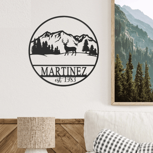 A circular wall art piece featuring a mountain landscape with a deer silhouette and the text \“MARTINEZ est 1983\“. It is hung above a decorative lamp and plaid pillow.