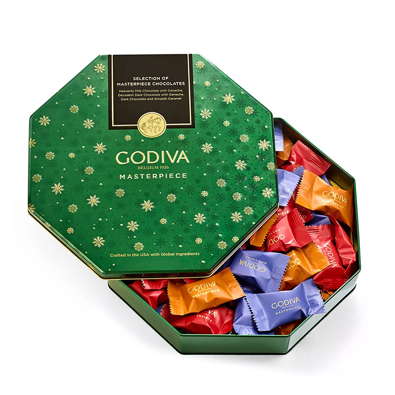 Godiva Masterpiece chocolates are presented in a green, snowflake-patterned hexagonal tin. The assortment includes milk chocolate with ganache, dark chocolate with ganache, and dark chocolate with smooth caramel, wrapped in colorful packaging.