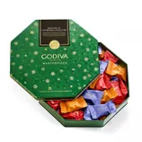 Godiva Masterpiece chocolates are presented in a green, snowflake-patterned hexagonal tin. The assortment includes milk chocolate with ganache, dark chocolate with ganache, and dark chocolate with smooth caramel, wrapped in colorful packaging.