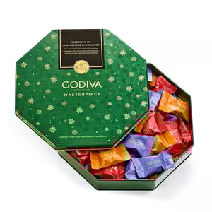 Godiva Masterpiece chocolates are presented in a green, snowflake-patterned hexagonal tin. The assortment includes milk chocolate with ganache, dark chocolate with ganache, and dark chocolate with smooth caramel, wrapped in colorful packaging.